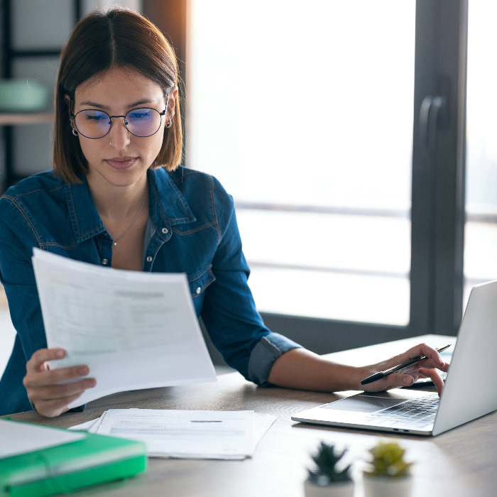 Woman wearing glasses while reviewing a paper and typing on a laptop