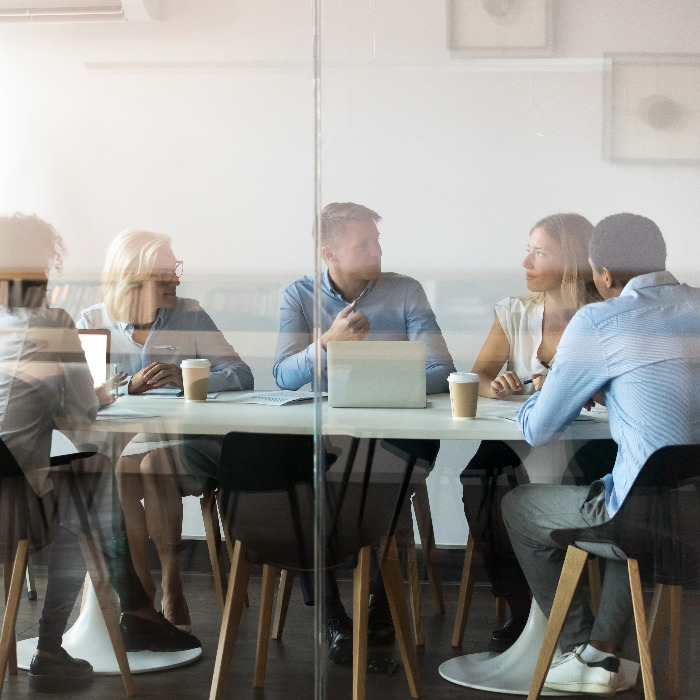 From behind a glass wall, five people sitting and discussing at a conference room table