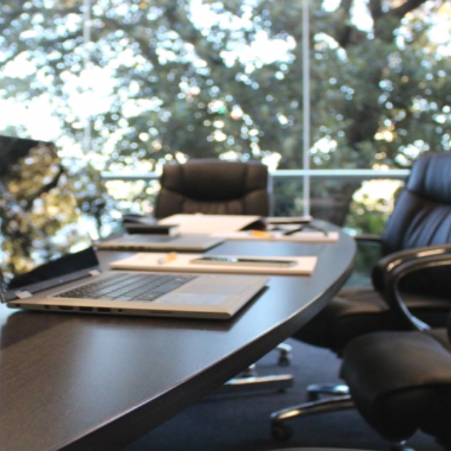 A laptop open on a long conference room table with a large window with trees outside in the backgroun