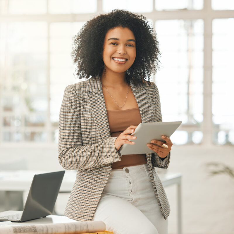 Woman in a houndstooth jacket smiling and holding a tablet while sitting on the edge of a desk