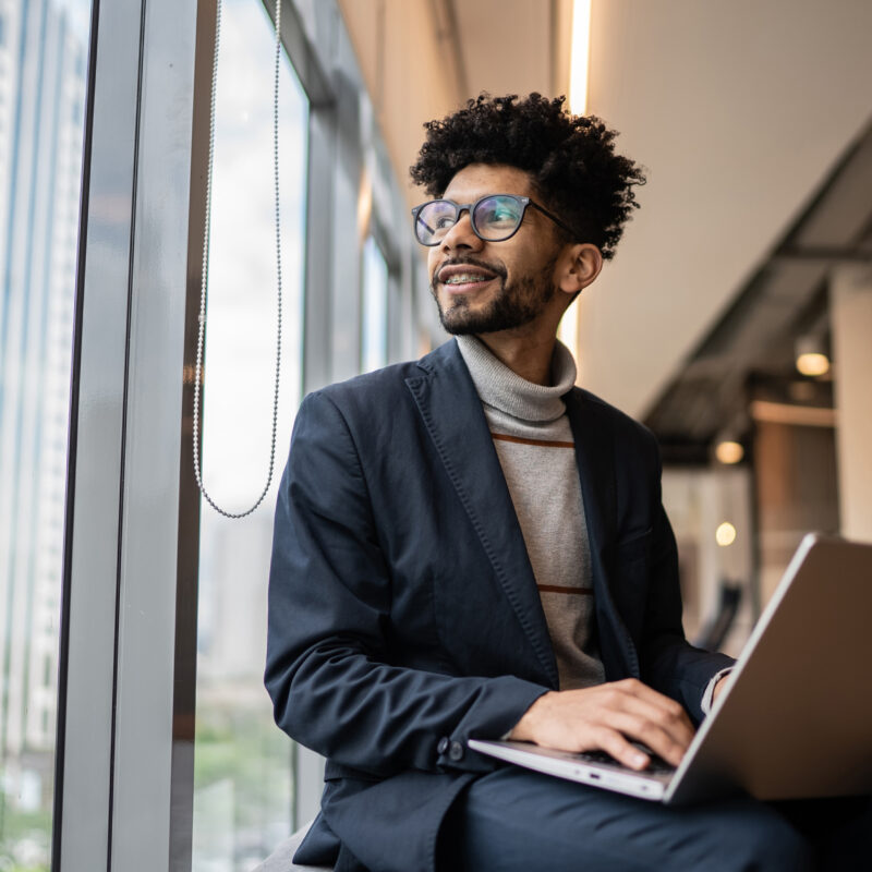 A man smiling and looking off to the side out the window with his hands on a keyboard laptop balanced on his lap