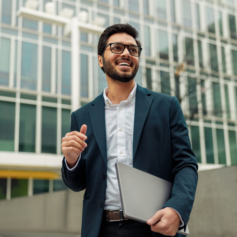 A man smiling looking off in the distance carrying a closed laptop and walking